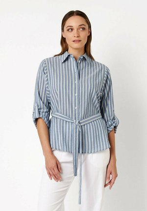 Frau trägt ein blau-weiß vertikal gestreiftes Hemd mit Knöpfen, aufgekrempelten Ärmeln und gebundenem Taillengürtel, kombiniert mit weißen Hosen.
