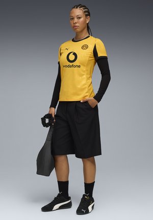 Gul fotbollströja med svarta detaljer, svarta shorts och svarta sneakers. Tröjan har logotyp och sponsor. Lätt och sportig design.