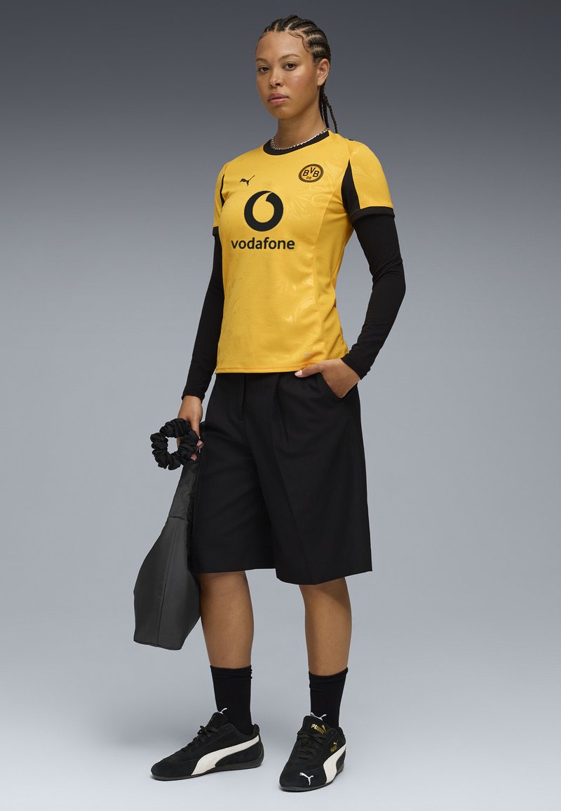 Maglia da calcio gialla con accenti neri, pantaloni neri e sneakers nere. La maglia presenta il logo e lo sponsor. Design leggero e sportivo.