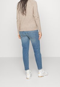 Persona con cabello largo trenzado, vistiendo un suéter beige, jeans azul, calcetas blancas y zapatillas blancas, de pie frente a un fondo liso.