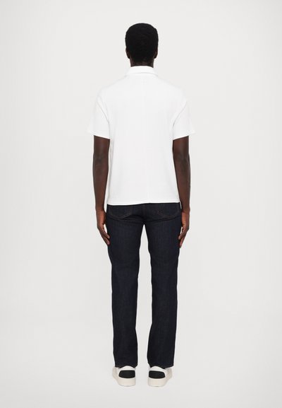 Chemise polo blanche avec un col, associée à un jean bleu foncé et des baskets blanches. La tenue présente des manches courtes et une coupe décontractée.
