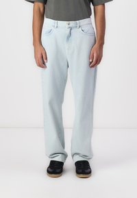 Light blue denim jeans with a relaxed fit, featuring a waistband with a button closure and front pockets. Worn with black sneakers.