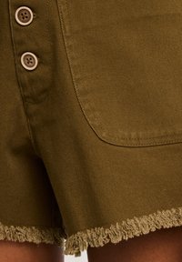 Alyvuogių spalvos šortai, pagaminti iš tvirtos medžiagos, turintys nusidėvėjusį kraštą, dvi dideles sagas ir matomą šoninę kišenę su tvarkingu užbaigimu.