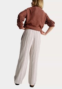 Pull en tricot couleur rouille avec des manches texturées, associé à un pantalon large rayé en blanc et rose clair. Chaussures noires visibles.