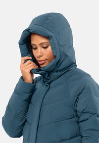 Giacca a vento in teal profondo, dotata di un ampio cappuccio regolabile, motivo trapuntato, chiusura con zip e una texture morbida e leggera.