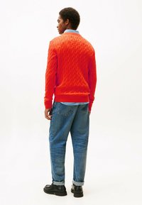 Maglione rosso a maglia grossa con scollatura rotonda, abbinato a una camicia azzurra con colletto, jeans denim blu larghi e scarpe nere.
