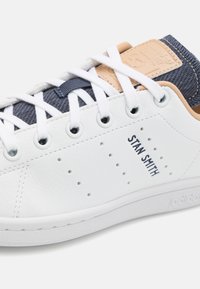 Valkoiset nahkakengät, joissa on teksturoitu tummansininen kangasvarsi ja vaaleanruskeat yksityiskohdat. Ominaisuuksiin kuuluu rei'itetty yksityiskohtaus ja "STAN SMITH" -brändäys.