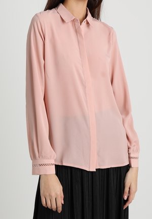 Vrouw draagt een lichtroze blouse met lange mouwen en knoopmanchetten en een zwarte geplooide rok, staand tegen een effen achtergrond.