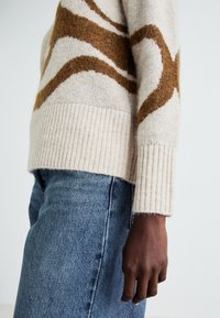Lysbeige genser med brune bølgemønstre, ribbestrikkede mansjetter og hem; brukt over blå denimjeans. Myk tekstur, uformell design.