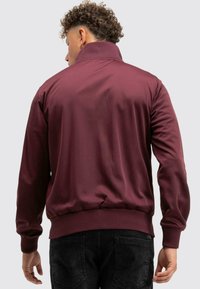 Burgundi színű, cipzáros dzseki magas gallérral, bordázott mandzsettákkal és aljjal. Simává anyag, testhezálló fazon és letisztult vonalak jellemzik.