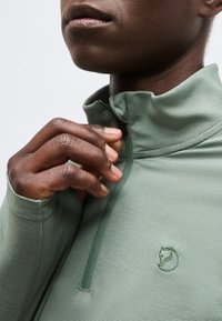 Tumšādains cilvēks aizvelk gaiši zaļu teksturētu jaku, kurai uz krūšu daļas ir neliels lapsas galvas logo.