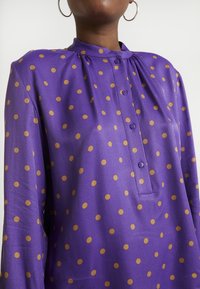 Blusa morada hecha de tela suave con lunares circulares marrones, con un cuello mandarín y una tapeta de botones.