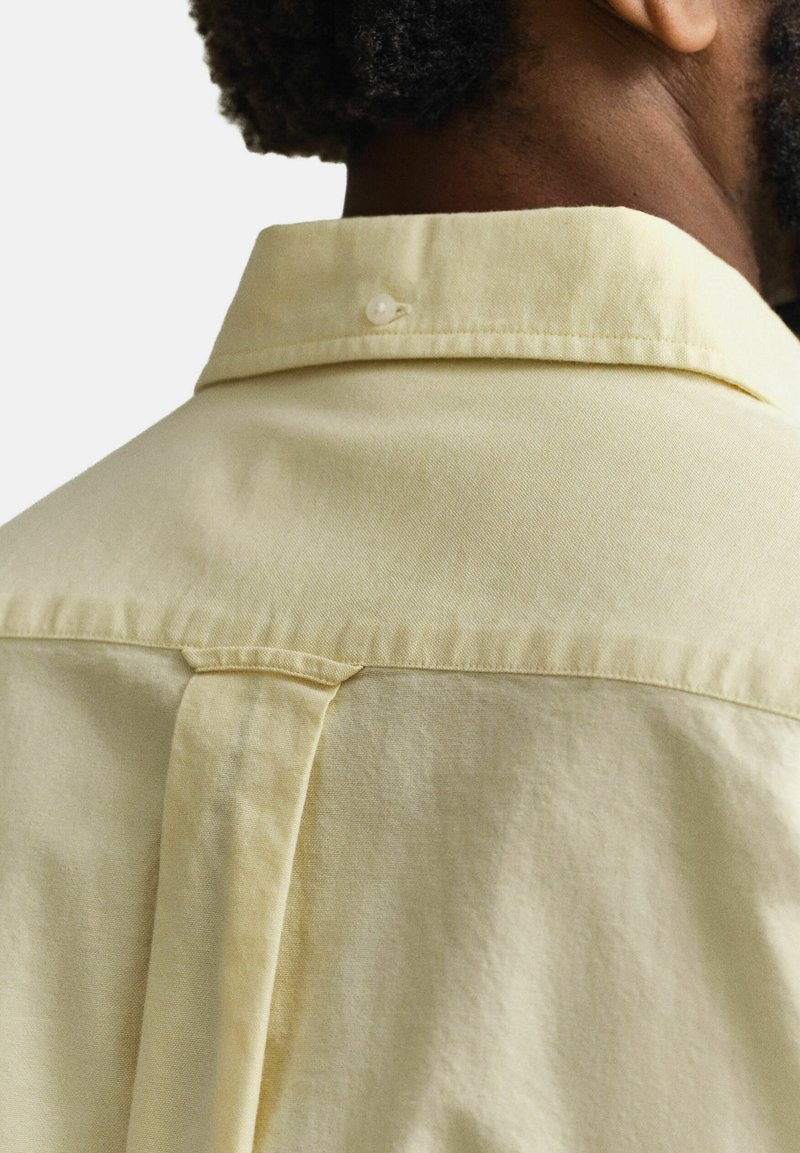 Primer plano de la parte superior de la espalda de una persona que lleva una camisa de botones de color amarillo pálido con una trabilla abotonada en el cuello y un pliegue en el centro de la espalda.