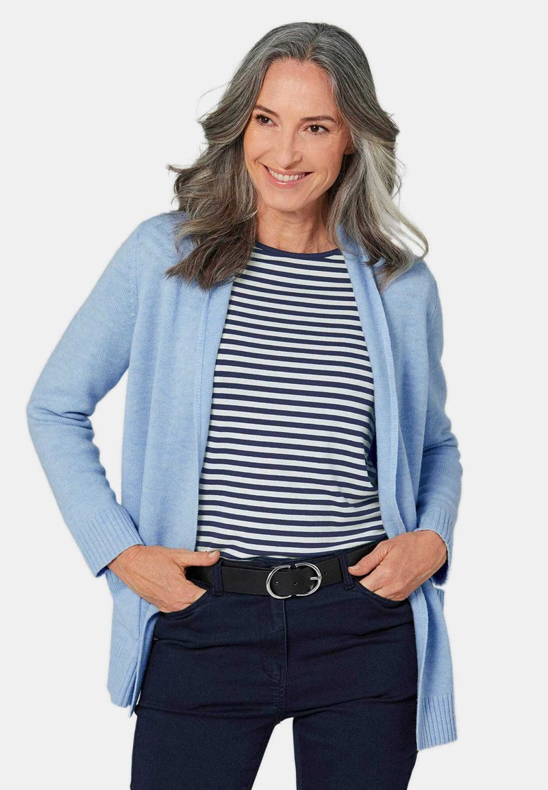 Cardigan bleu clair avec devant ouvert, poignets côtelés. Chemise rayée bleu marine et blanche en dessous, accompagnée de pantalons sombres et d'une ceinture noire.