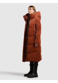 Mujer con abrigo largo acolchado de color óxido con cuello alto y capucha, botas negras, de perfil sobre un fondo liso.