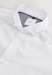 Chemise blanche à boutons avec tissu texturé, col classique et détail intérieur du col à motif avec une étiquette de marque visible "NEXT".