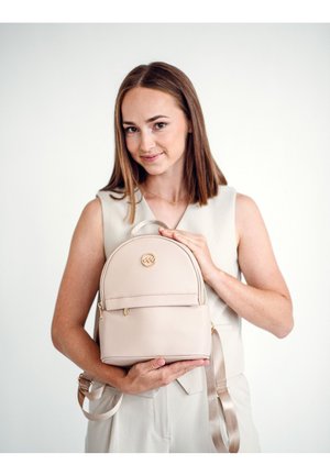 Beige faux leather backpack with rounded top, front zipper pocket, and gold logo. Features adjustable straps and a sleek, smooth texture.