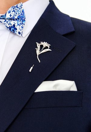 Granatowa marynarka z srebrną broszką w kształcie kwiatu na klapie, białą poszetką i niebieską muchą w kwiatowy wzór z wyszukanymi motywami roślinnymi.