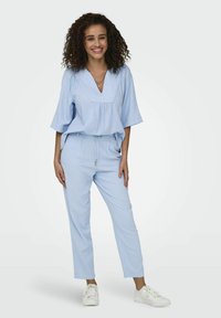 Jumpsuit azzurro chiaro con scollatura a V, maniche corte, vita elastica e gambe tapered, abbinato a sneakers bianche. Tessuto morbido al tatto.