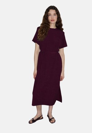 Frau mit langen lockigen Haaren, die ein dunkelviolettes Midikleid mit kurzen Ärmeln und schwarze offene Sandalen trägt, steht vor einem einfarbigen Hintergrund.