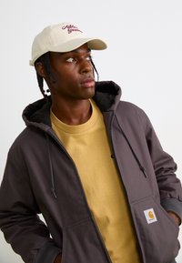 Dark gray hooded jacket with a front pocket, layered over a yellow shirt. A light-colored cap features "Athlete Garments" in red script.