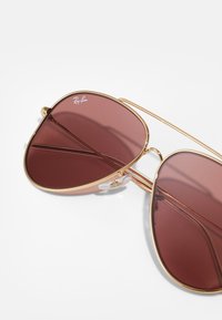 Sluneční brýle Ray-Ban se zlatým kovovým rámem a tmavě červenými čočkami. Čočky mají tvar kapky se průhlednými nosními podložkami a viditelným logem.