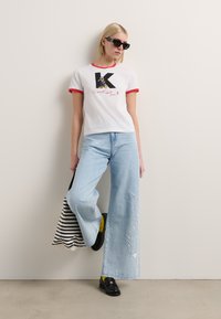 Weißes T-Shirt mit rotem Ringkragen, versehen mit einem Grafikdesign eines Charakters und „K.“ Hellblaue weite Jeans mit weißen bestickten Details. Schwarze Schuhe.