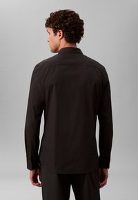 Čierna košeľa s dlhým rukávom, hladkou textúrou, špicatým golierom a zapínaním na gombíky na manžetách. Strihaná priliehavo s lemom, ktorý jemne zaobľuje vzadu.