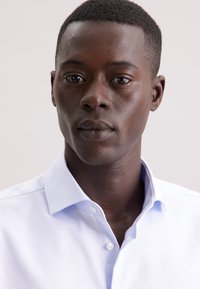 Un giovane uomo di colore con capelli corti, che indossa una camicia azzurro chiaro con bottoni, guarda direttamente con espressione neutra su uno sfondo chiaro e semplice.