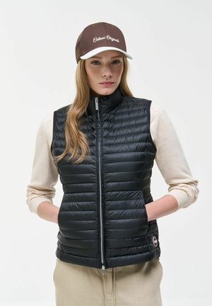 Giovane donna che indossa un berretto marrone e bianco, giacca smanicata trapuntata nera sopra una maglia beige a maniche lunghe, mani nelle tasche, sfondo neutro.