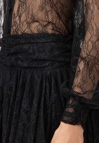 Robe noire en dentelle avec détails transparents, présentant un tissu texturé, une taille froncée et des manches à volants. Motif floral élégant sur toute la surface.