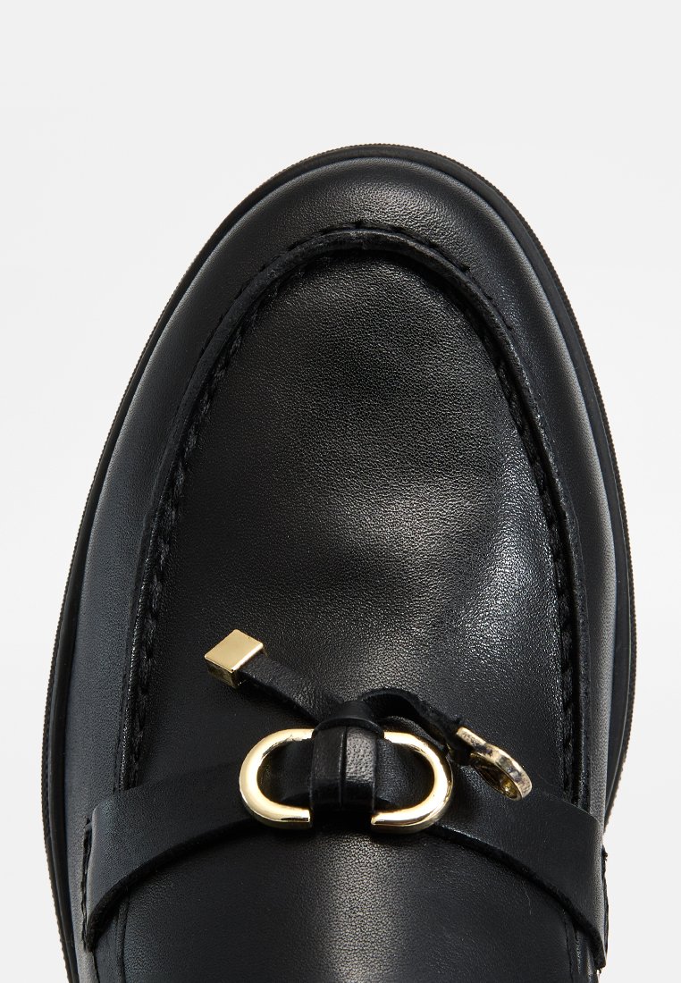 Mocassin en cuir noir avec un bout arrondi, featuring une boucle en métal doré et un détail de sangle en cuir sur le devant.