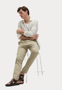 Beige chino's, wit tanktop en een lichtgrijze open overhemd. Bruine sandalen met bandjes. Model zit op een witte kruk.