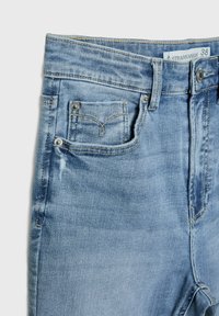 Jean en denim bleu clair en gros plan montrant la poche avant, petite poche à monnaie avec rivets, passants de ceinture, bouton et étiquette de marque.