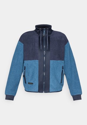 Fliisjakki, millel on värviblokeerimise disain sinise ja tumesinise toonides, kõrge krae, kaks tõmblukuga taskut ja reguleeritavad nöörid.