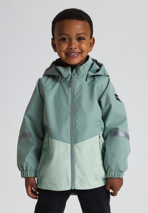 Lachend kind dat een tweekleurige groene capuchonjas draagt met een ritssluiting aan de voorkant en reflecterende strepen op de mouwen, staand tegen een effen achtergrond.