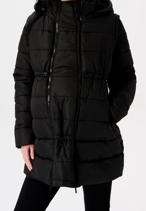 Femme portant un manteau d'hiver noir matelassé avec capuche, fermeture éclair devant et mains dans les poches sur un fond uni.