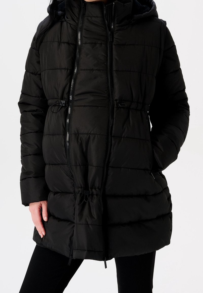 Femme portant un manteau d'hiver noir matelassé avec capuche, fermeture éclair devant et mains dans les poches sur un fond uni.