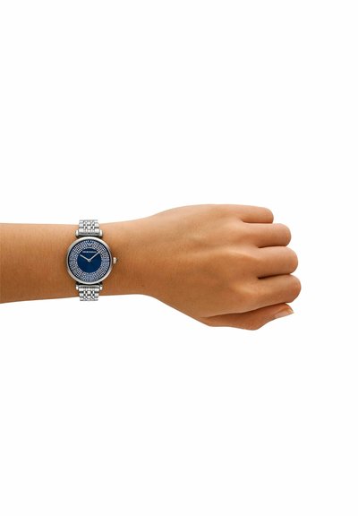 Pols met een zilverkleurig metalen schakelhorloge met een blauwe wijzerplaat en kristallen details, dat de tijd 10:10 aangeeft.