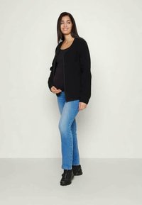 Cardigan preto por cima de uma blusa preta, combinado com jeans azul claro e sapatos pretos. O cardigan tem um design simples e está aberto na frente.