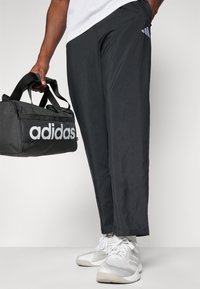 Mustad mustad püksid elastse pehmendusega ja hall Adidas logo. Inimene hoiab musta spordikotti valge Adidas brändinguga. Valged tossud.