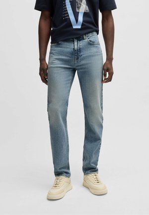 Man wearing faded blue straight-leg jeans, navy graphic t-shirt, and beige lace-up sneakers standing against plain background.