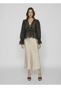 Svart lace-blus med utskurna mönster, djup V-ringning och puffärmar, i kombination med en slät, ljusbeige satinskjol och svarta klackskor.