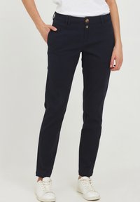Mørkeblå slim-fit bukser med to frontlommer, knap lukning og dobbelt knaplukning. Båret med en hvid top og hvide sneakers.