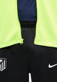 Gros plan d'une personne portant un pantalon noir Nike avec le logo de l'Atlético Madrid et une veste jaune néon partiellement zippée sur un t-shirt sombre.
