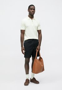 Polo à fermeture éclair vert clair, short noir, sac fourre-tout en cuir marron, baskets marron et chaussettes blanches. Simplicité dans le design et le contraste des couleurs.