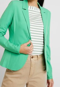 Blazer vert au design ajusté, avec un col cranté et une texture lisse. Porté sur une chemise rayée et un pantalon marron clair.
