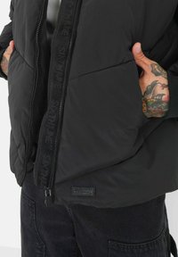 Svart puffer-jakke med quiltet tekstur, med glidelås foran, merket glidelåsbånd og en liten logotagg på kanten.