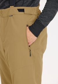 Beige geïsoleerde broek met een elastische tailleband, zijrits zak en een gladde textuur. Een contrasterend zwart langemouwen shirt is zichtbaar.