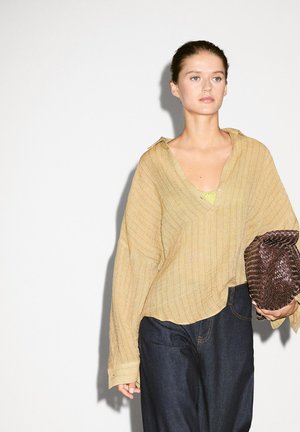 Camicia oversize gialla chiara con texture, scollatura a V e maniche larghe; pantaloni in denim blu navy; porta una pochette intrecciata marrone.